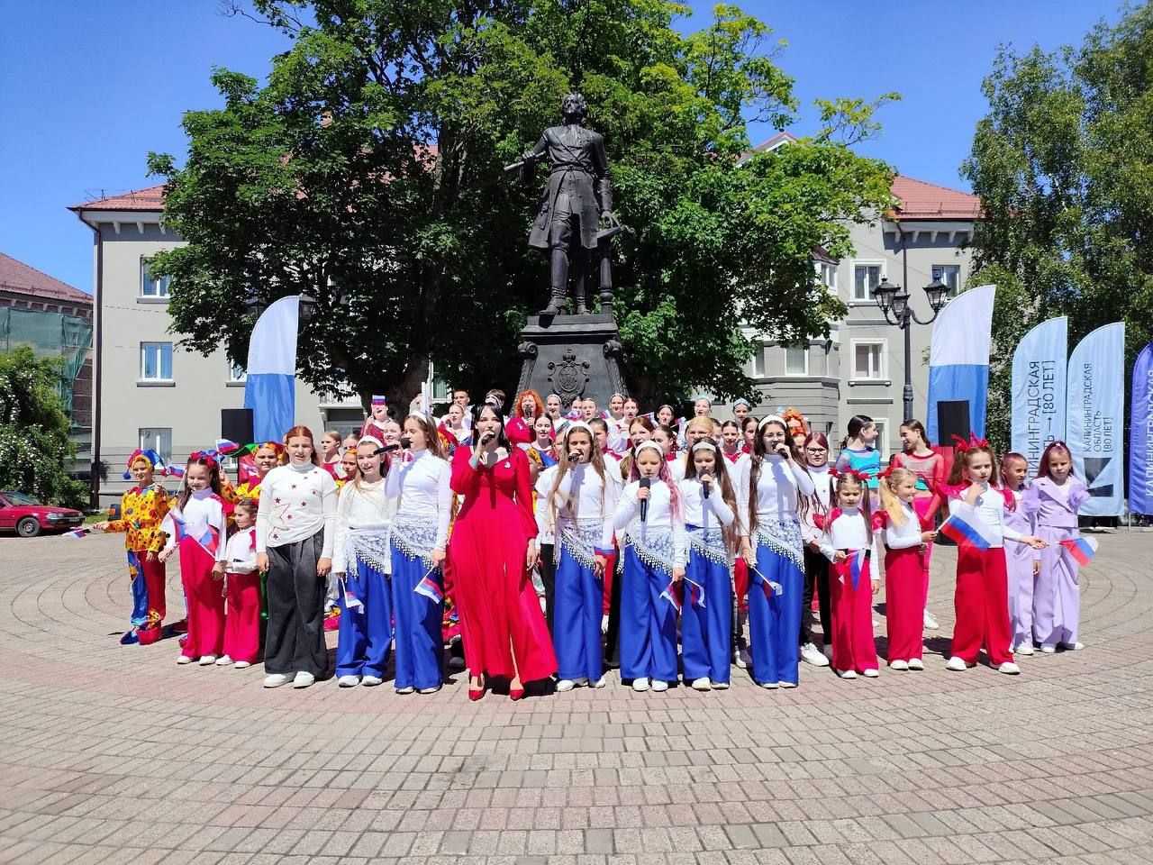От спортивных площадок у Верхнего озера наш праздничный маршрут переносит нас в самый западный город страны — Балтийск. Здесь, у величественного памятника Петру I, развернулось масштабное торжество, запечатленное на этом снимке. На площади, украшенной флагами и баннерами в честь 80-летия Калининградской области, сотни горожан наблюдали за яркой программой. Центральным событием стали выступления молодых артистов Культурно-молодежного центра, вышедших в колоритных костюмах патриотических расцветок. Помимо концерта, праздник у нас в приморском городе включил в себя вручение паспортов юным жителям и торжественное чествование героев-водолазов центра «Лидер», которые обезвредили тонны опасных боеприпасов в наших водах.